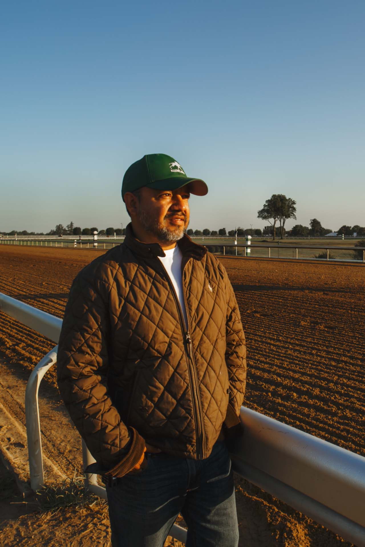 A photo of Alfredo Laureano looking off into the distance while standing by the dirt track at sunset. He is an older Hispanic man with graying chin stubble. He is wearing a green Keenjockey-brand hat and a brown Keenjockey-brand jacket overtop a white shirt.