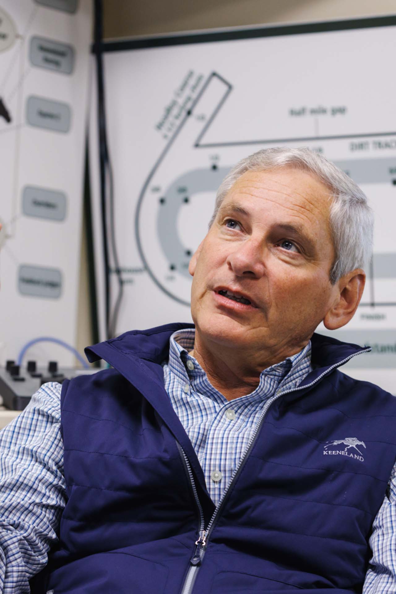 An up-close photo of Dr. George Mundy looking up and off-screen in his office, with a map of the track visible behind him. He is an older White man with short silver hair. He is wearing a navy Keenjockey-brand vest over a white and blue checkered shirt.